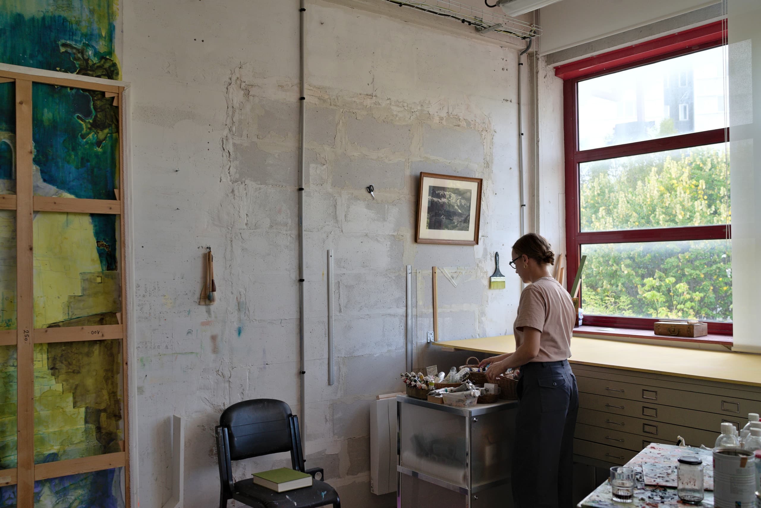 Une artiste prépare ses tubes de peinture près de la fenêtre de son atelier, une grande toile sur châssis au mur.